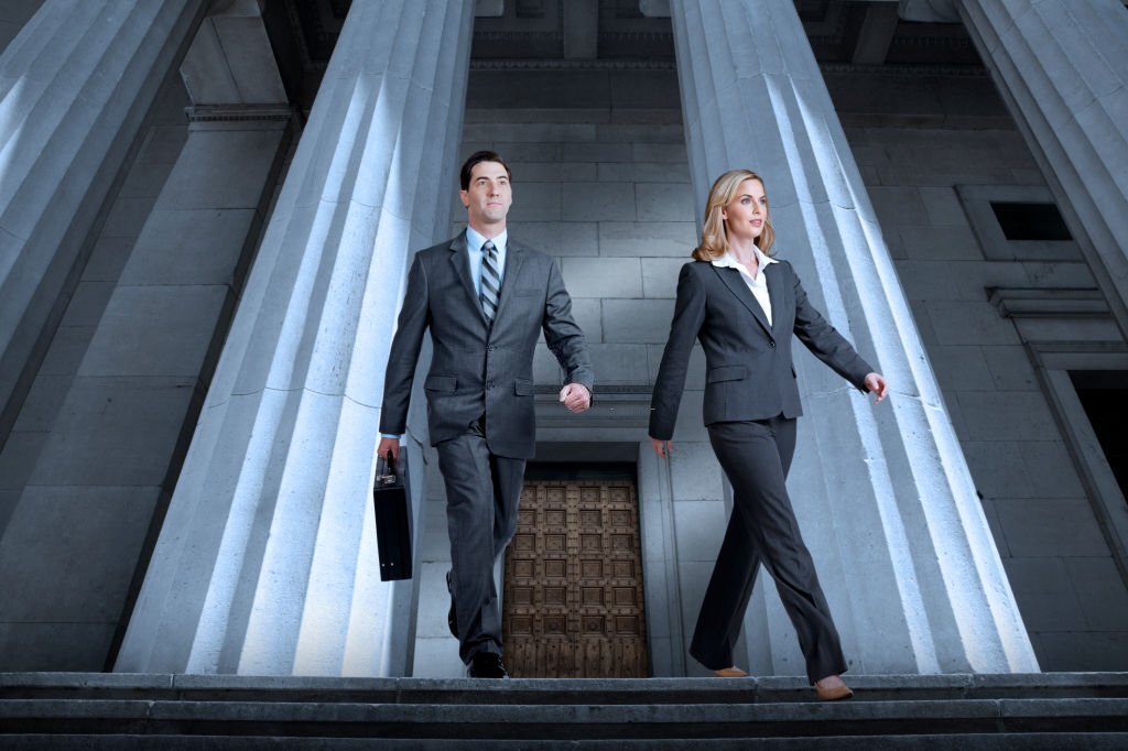 Two lawyers, one a man and the other a woman, dressed in suits walk briskly away from a column supported courthouse as they are about to walk down the steps as the leave the building.