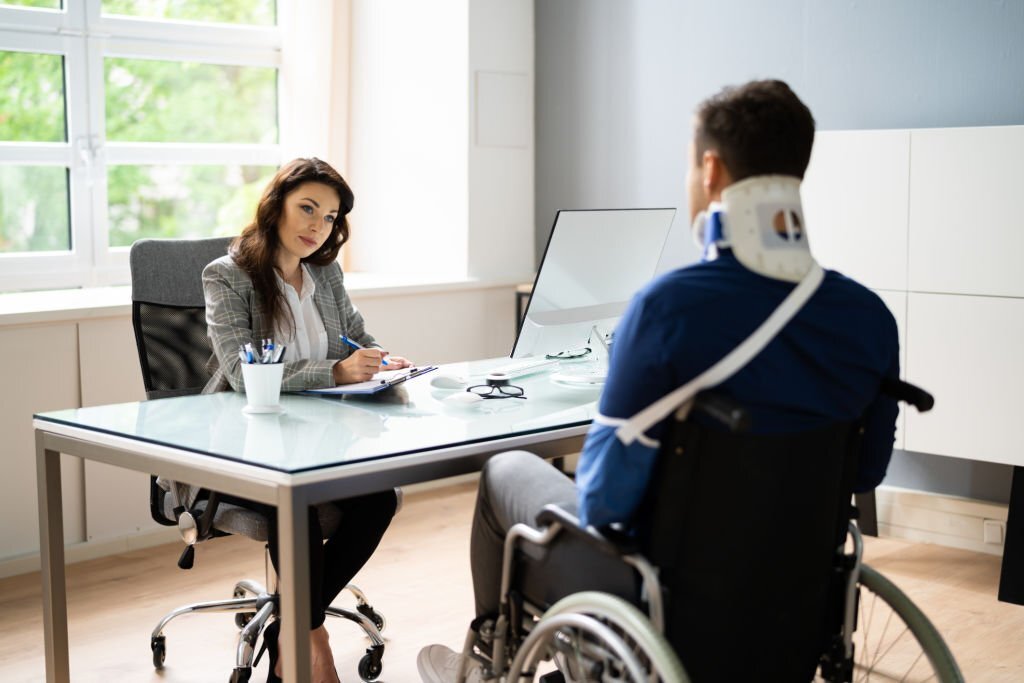 Worker Injury And Disability Compensation. Social Security Claim- A woman sitting in an office chair engaged in conversation with a man in wheelchairs.